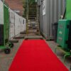 A Heavy-Duty PVC Vinyl Roll in bright red leads to a metal outdoor staircase, surrounded by construction barriers, green trolleys, and equipment cases for an industrial, utilitarian setting.