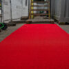 A Heavy-Duty PVC Vinyl Roll in bright red runs along an outdoor path, flanked by construction materials, green trolleys, and a wooden staircase leading to a raised platform.