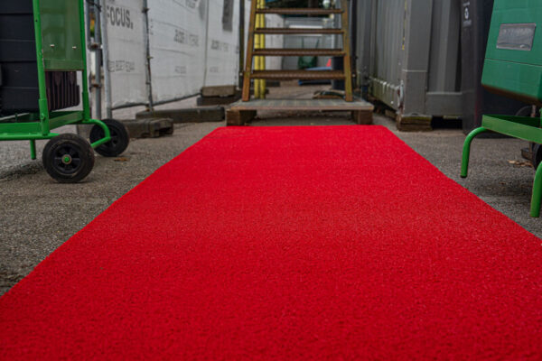 A Heavy-Duty PVC Vinyl Roll in bright red runs along an outdoor path, flanked by construction materials, green trolleys, and a wooden staircase leading to a raised platform.