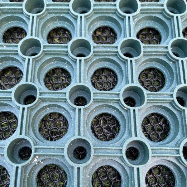 Close-up of the Green Grass Mat showing its textured green rubber surface with a grid of large circular holes, small corner circles, and a dark mesh-like layer beneath.