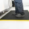 A person wearing dark shoes and trousers stands on a Safety Cross-Ribbed Mat with a yellow border in a clean indoor space.