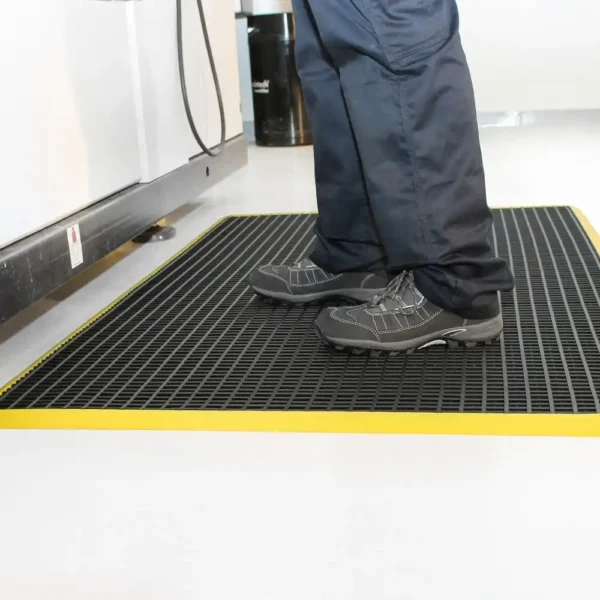 A person wearing dark shoes and trousers stands on a Safety Cross-Ribbed Mat with a yellow border in a clean indoor space.