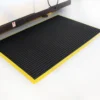 A Safety Cross-Ribbed Mat with a black surface and yellow border is placed on a white floor beside a brown desk or workbench.