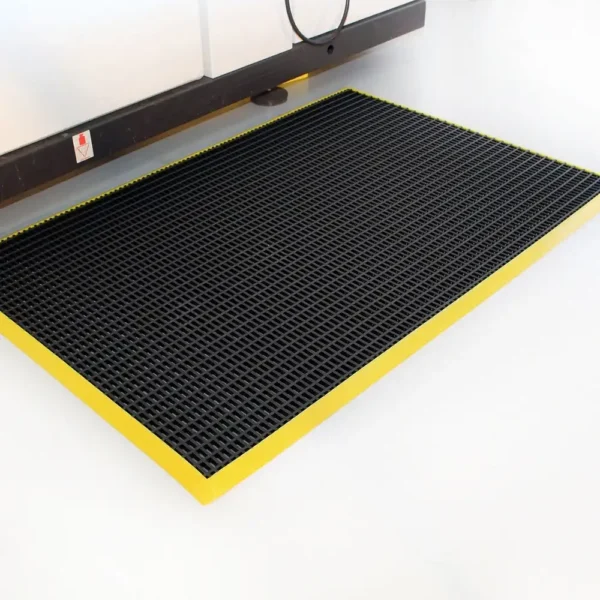 A Safety Cross-Ribbed Mat with a black surface and yellow border is placed on a white floor beside a brown desk or workbench.