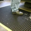 A person in jeans and black work shoes stands on a Safety Cross-Ribbed Mat with a yellow border, which has metal shavings and liquid visible on its black surface.