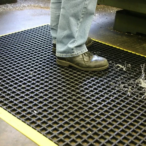 A person in jeans and black work shoes stands on a Safety Cross-Ribbed Mat with a yellow border, which has metal shavings and liquid visible on its black surface.