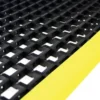 Close-up of the Safety Cross-Ribbed Mat, a sturdy black industrial grid with square openings and a bright yellow edge stripe, designed for safe flooring in work areas.