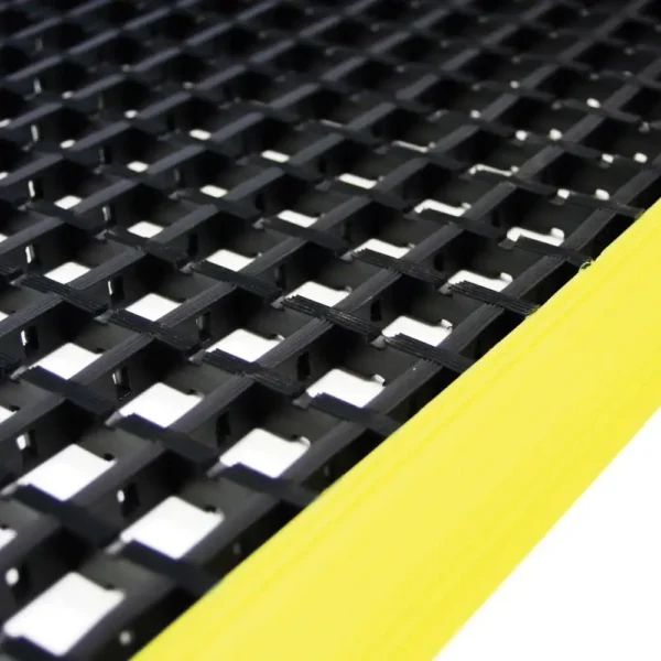 Close-up of the Safety Cross-Ribbed Mat, a sturdy black industrial grid with square openings and a bright yellow edge stripe, designed for safe flooring in work areas.