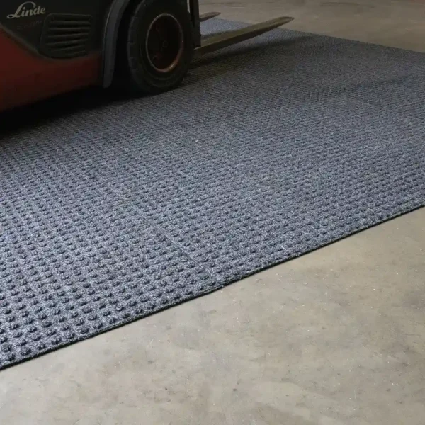 A forklift wheel rests on the Heavy-Duty Heavy Traffic Doorway Mat, a textured grey mat placed on a smooth concrete floor in an industrial or warehouse environment.