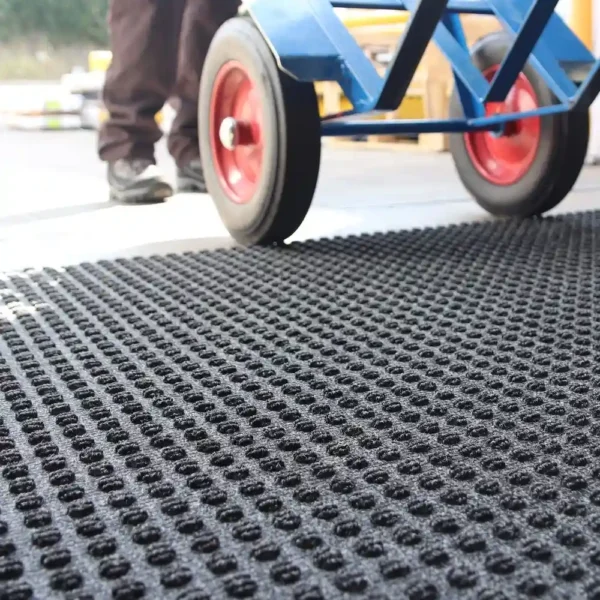 Close-up of the Heavy-Duty Heavy Traffic Doorway Mat, featuring a textured, perforated black rubber surface. In the background, a person stands beside a blue sack truck with red wheels on a concrete floor.