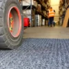Close-up of a forklift tyre on the Heavy-Duty Heavy Traffic Doorway Mat in a warehouse, with shelves on both sides and a worker in an orange safety vest walking away in the background.