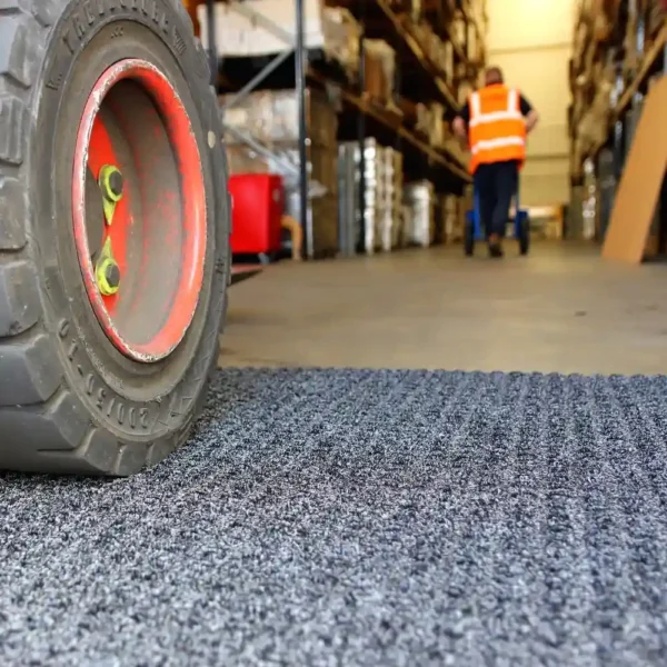 Close-up of a forklift tyre on the Heavy-Duty Heavy Traffic Doorway Mat in a warehouse, with shelves on both sides and a worker in an orange safety vest walking away in the background.