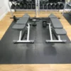 Two adjustable weight benches are placed on an interlocking gym mat, facing each other with a barbell, dumbbells, and weight plates between them. A rack of dumbbells is in the background on a wooden floor.