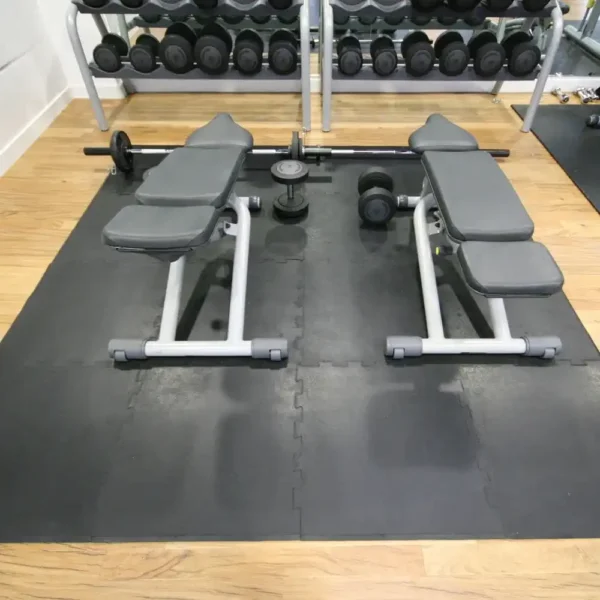 Two adjustable weight benches are placed on an interlocking gym mat, facing each other with a barbell, dumbbells, and weight plates between them. A rack of dumbbells is in the background on a wooden floor.