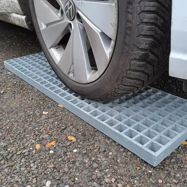 A car tyre rests on Waffle Boards, a rectangular metal grid platform on gravel, with part of the wheel and car body visible.