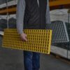 A person in a grey jumper and black waistcoat stands in an industrial warehouse, holding yellow and grey Waffle Boards—grated panels designed for versatile industrial use.