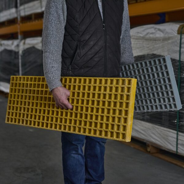 A person in a grey jumper and black waistcoat stands in an industrial warehouse, holding yellow and grey Waffle Boards—grated panels designed for versatile industrial use.