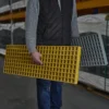 A person in a grey jumper and black waistcoat holds yellow and grey Waffle Boards while standing in an industrial warehouse.