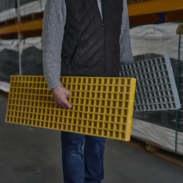 A person in a grey jumper and black waistcoat holds yellow and grey Waffle Boards while standing in an industrial warehouse.