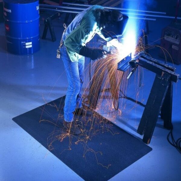 A person in protective gear welds metal at a workbench, with bright blue sparks landing on a Welding Anti-Fatigue Mat placed on the workshop floor.