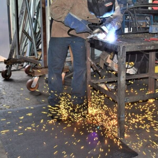 In a workshop, a person in protective gear welds metal at a workbench as bright sparks fly, standing on the Welding Anti-Fatigue Mat for comfort and safety.