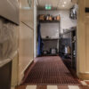 A narrow kitchen corridor features a red Honeycomb Kitchen Mat, stainless steel shelves with pots and pans, an emergency exit sign above the door, plus a bin to the left and worktop to the right.