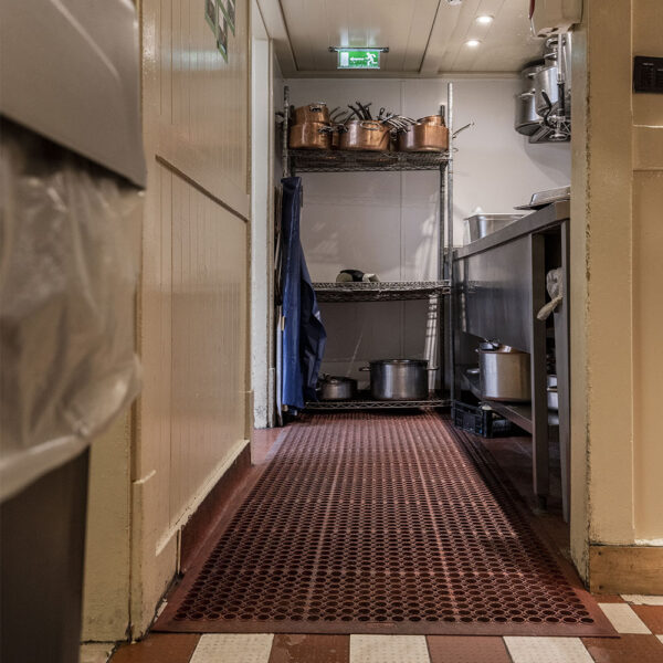 A narrow kitchen corridor features a red Honeycomb Kitchen Mat, stainless steel shelves with pots and pans, an emergency exit sign above the door, plus a bin to the left and worktop to the right.