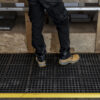 A person in black work trousers and tan safety boots stands on an Interlocking Anti-Fatigue Mat with holes, placed in front of a wooden workbench. A yellow safety line marks the mat’s edge.