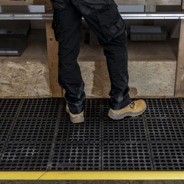 A person in black work trousers and tan safety boots stands on an Interlocking Anti-Fatigue Mat with holes, placed in front of a wooden workbench. A yellow safety line marks the mat’s edge.