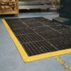 An Interlocking Anti-Fatigue Mat with black and yellow edges lies on a blue workshop floor, surrounded by wood shavings and industrial equipment.