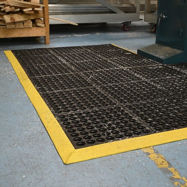 An Interlocking Anti-Fatigue Mat with black and yellow edges lies on a blue workshop floor, surrounded by wood shavings and industrial equipment.