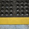 A close-up of Interlocking Anti-Fatigue Mats with circular holes and wood shavings, separated by a seam and bordered at the bottom by a textured yellow strip on a worn blue floor.