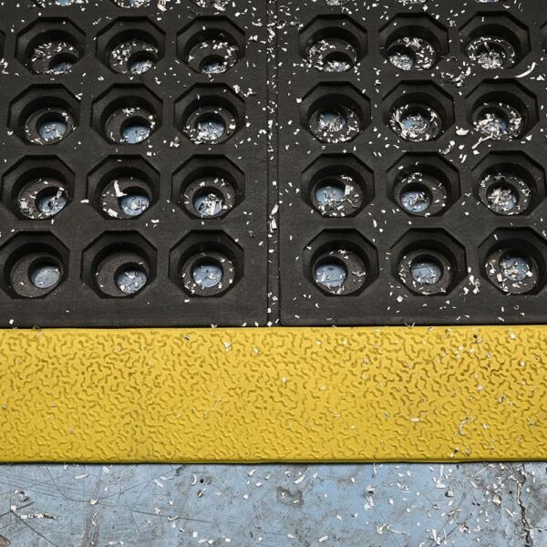 A close-up of Interlocking Anti-Fatigue Mats with circular holes and wood shavings, separated by a seam and bordered at the bottom by a textured yellow strip on a worn blue floor.