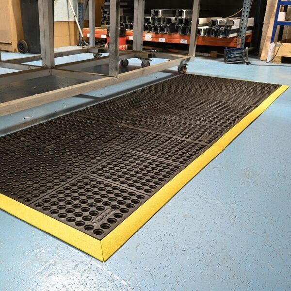 An Interlocking Anti-Fatigue Mat with yellow edges is positioned on a blue industrial floor next to metal workstations in a warehouse or factory setting.