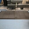 An Interlocking Solid Top Gym Mat with a grid pattern is placed on a blue workshop floor in front of industrial machinery, with metal swarf scattered nearby.