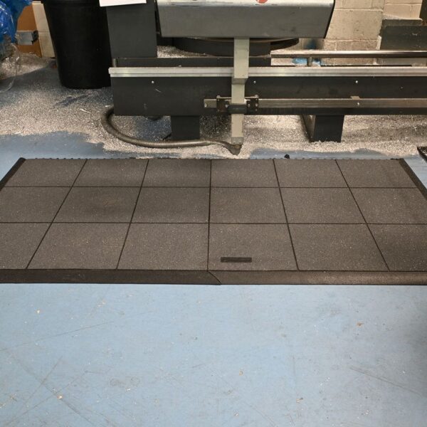 An Interlocking Solid Top Gym Mat with a grid pattern is placed on a blue workshop floor in front of industrial machinery, with metal swarf scattered nearby.
