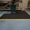 An Interlocking Solid Top Gym Mat with a yellow border is placed on a light blue industrial floor beneath a machine in a workshop, with shelving and equipment seen in the background.