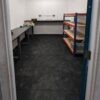 A small, empty room with dark tiled Interlocking Solid Top Gym Mats, two workbenches along the left and back walls, orange metal shelving with wooden shelves on the right, and bright white walls with overhead lighting.