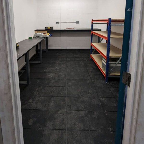 A small, empty room with dark tiled Interlocking Solid Top Gym Mats, two workbenches along the left and back walls, orange metal shelving with wooden shelves on the right, and bright white walls with overhead lighting.