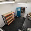 A clean, organised workshop with an Interlocking Solid Top Gym Mat floor, a blue door, empty three-tier metal shelves, worktops with assorted items, and a view into a cluttered adjoining room.
