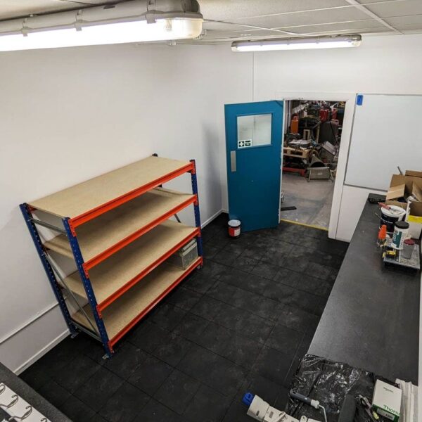A clean, organised workshop with an Interlocking Solid Top Gym Mat floor, a blue door, empty three-tier metal shelves, worktops with assorted items, and a view into a cluttered adjoining room.