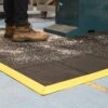 A person in brown work boots stands on an Interlocking Solid Top Gym Mat with yellow edges and a black surface, beside a machine. Wood or metal shavings are scattered across the mat and nearby floor.