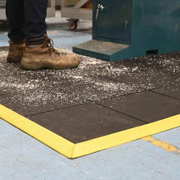 A person in brown work boots stands on an Interlocking Solid Top Gym Mat with yellow edges and a black surface, beside a machine. Wood or metal shavings are scattered across the mat and nearby floor.