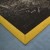 A close-up of the Interlocking Solid Top Gym Mat, featuring a black rubber surface with a textured yellow edge on a blue floor. White debris and shavings are scattered across the mat.