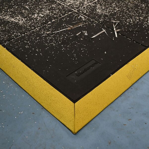 A close-up of the Interlocking Solid Top Gym Mat, featuring a black rubber surface with a textured yellow edge on a blue floor. White debris and shavings are scattered across the mat.