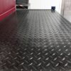 A close-up of Chequer Plate Rubber Matting in black, featuring a diamond pattern with some red and grey objects visible along the edge in the background.