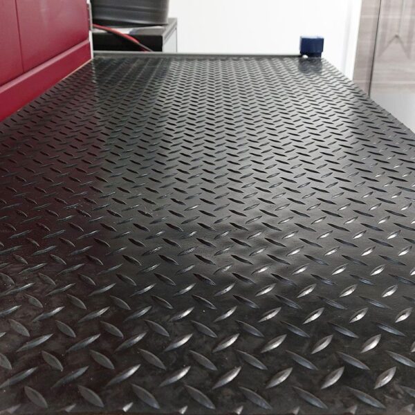 A close-up of Chequer Plate Rubber Matting in black, featuring a diamond pattern with some red and grey objects visible along the edge in the background.