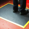 A person stands on the Air Cushioned Workshop Mat—a black anti-fatigue mat with raised bumps and a yellow border—placed on a red floor, showing only their lower legs and feet.