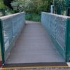 A small pedestrian bridge with solid top grating, featuring green railings and wire mesh sides, spans a gravel path surrounded by lush greenery and trees.