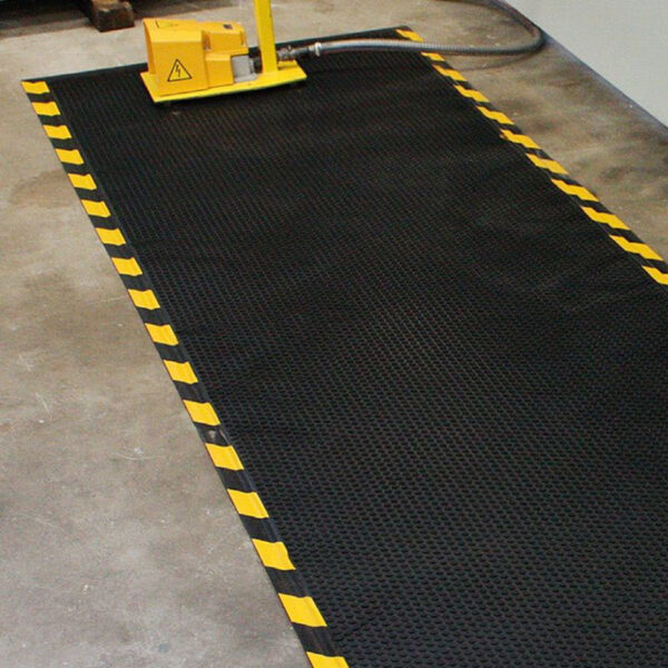A Foam Cushioned Industrial Mat with yellow and black striped edges is placed on a concrete floor. A yellow machine with a hose appears partially visible at the top of the image.